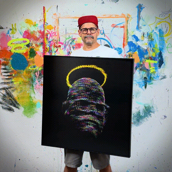 A man in a red hat and glasses stands in front of a colorful, abstract painted wall, holding a painting of a beaded head with a yellow halo on a black background.