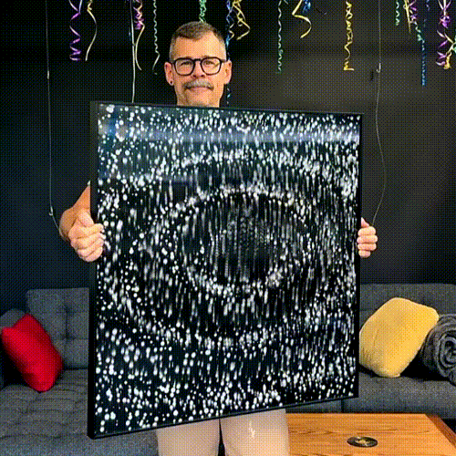 A smiling person with glasses holds a square artwork showing animated, sparkling white dots forming a swirling pattern. Party streamers hang from the ceiling and colorful cushions decorate a gray sofa in the background.
