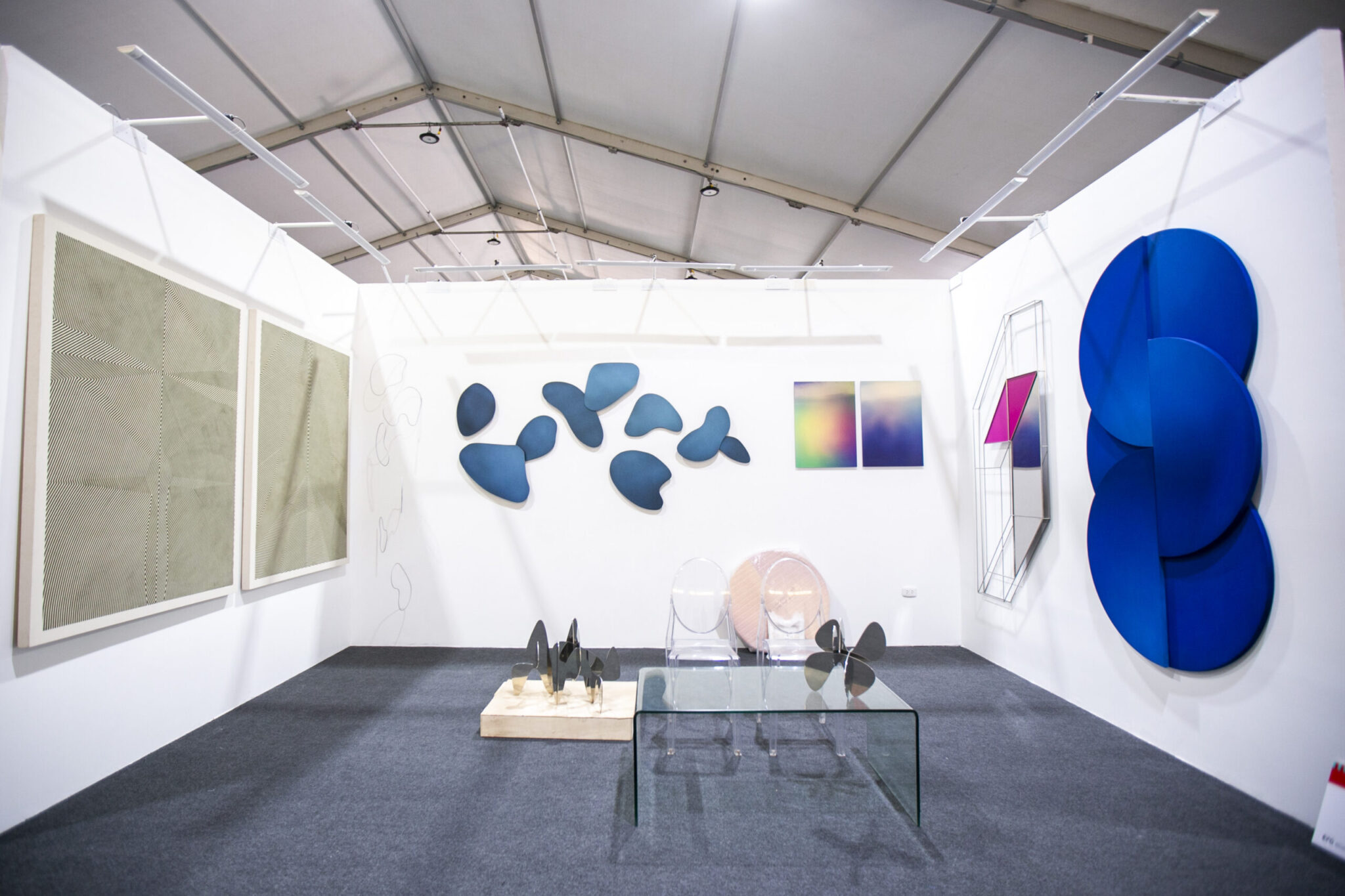 A modern art gallery with abstract paintings, blue wall sculptures, geometric art, and a glass table with acrylic chairs displayed on a dark carpet under a white ceiling.