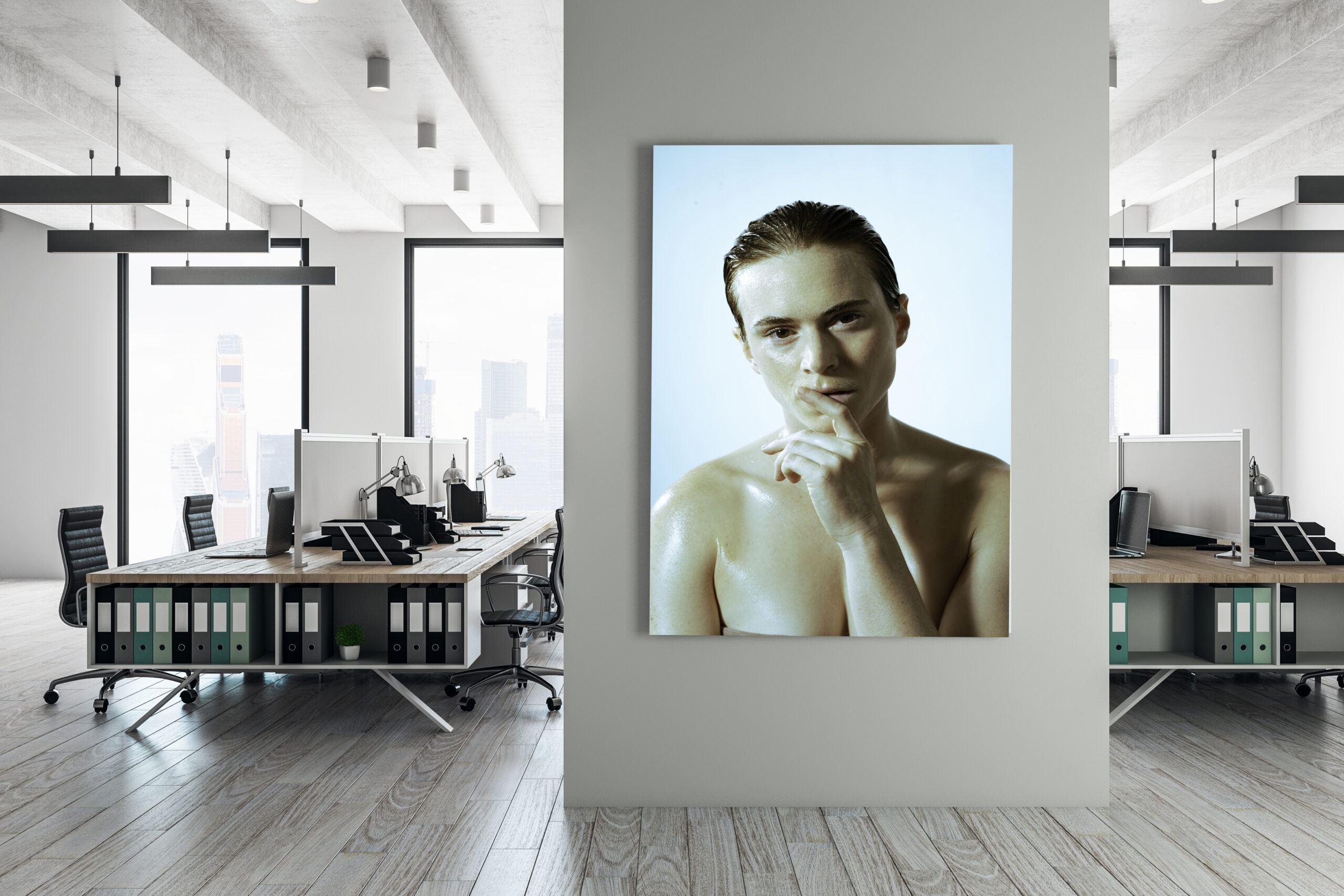 A modern office with desks, computers, and shelves, divided by a wall featuring a large, framed portrait of a person with wet hair and bare shoulders, touching their lips thoughtfully.