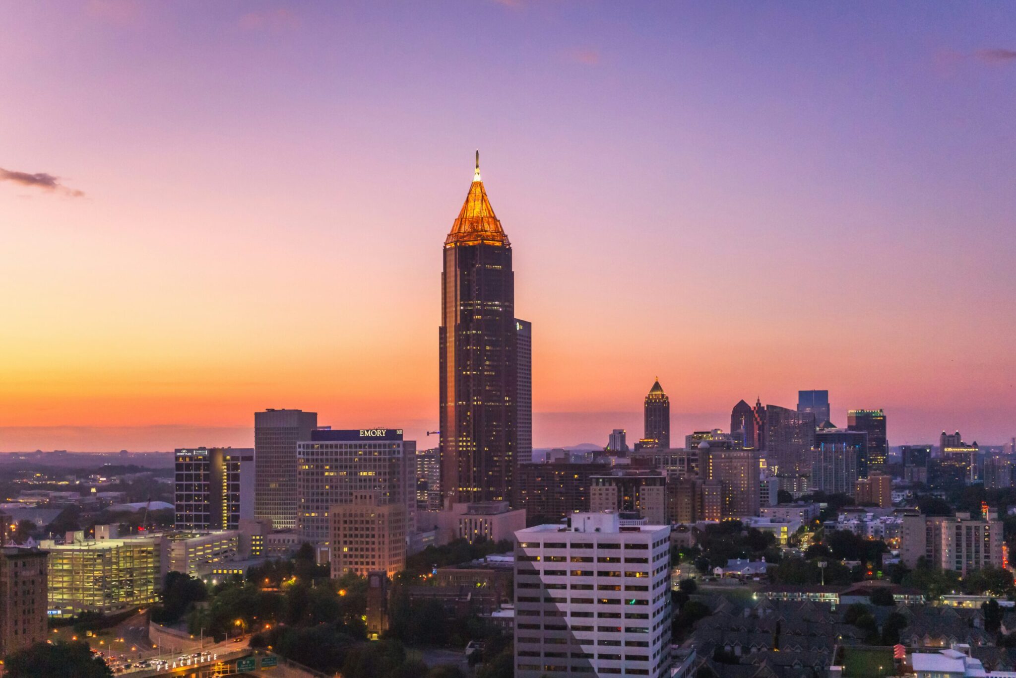 Atlanta city skyline at sunset, featuring a tall building with a gold triangular top, surrounded by other high-rise buildings, under a colorful orange and purple sky.