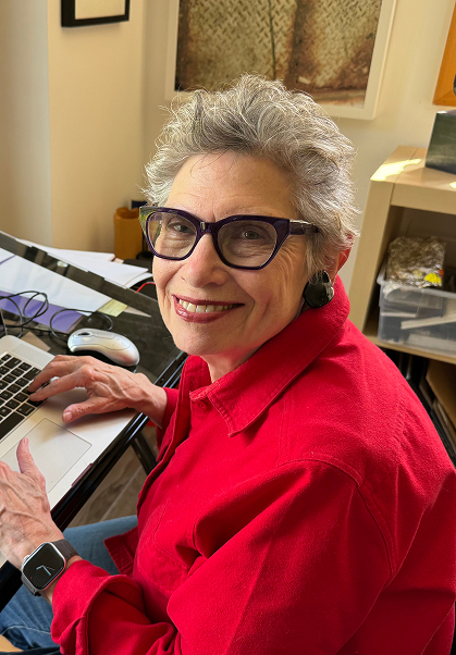 An older woman with short gray hair and glasses smiles at the camera while sitting at a desk with a laptop. She is wearing a bright red shirt and a smartwatch. Papers and office supplies are visible around her.
