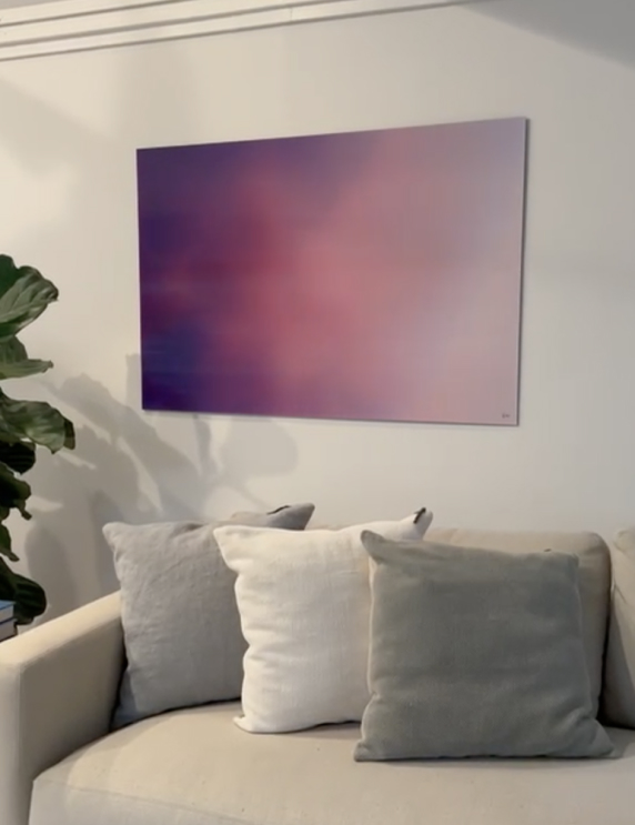 A modern living room with a beige sofa, three cushions in gray and white, a tall green plant, and an abstract purple and pink artwork hanging on the wall above the sofa.