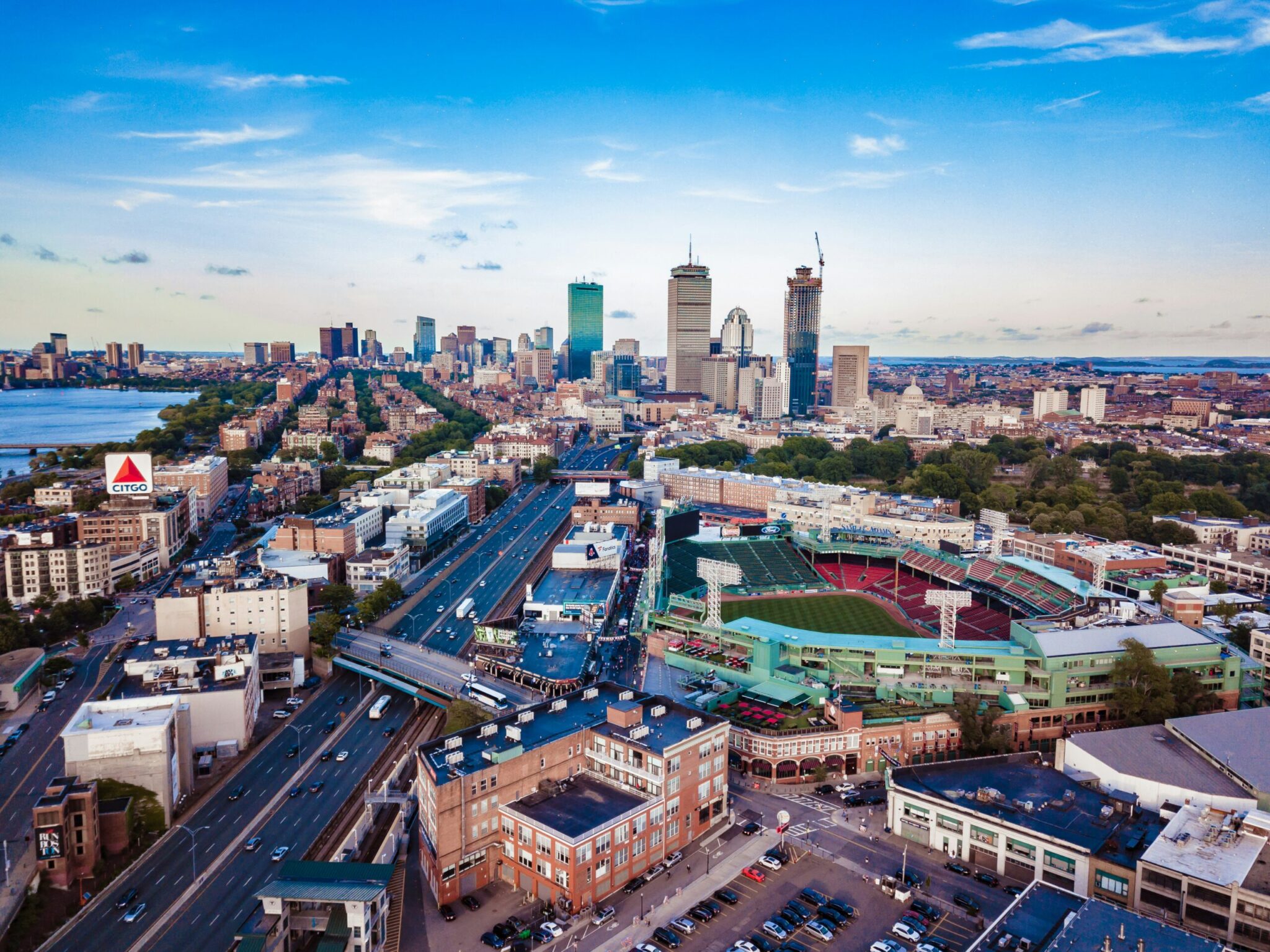 Aerial view of Boston featuring Fenway Park, busy roads, city buildings, and the Prudential and John Hancock towers in the distant skyline under a blue sky with scattered clouds.