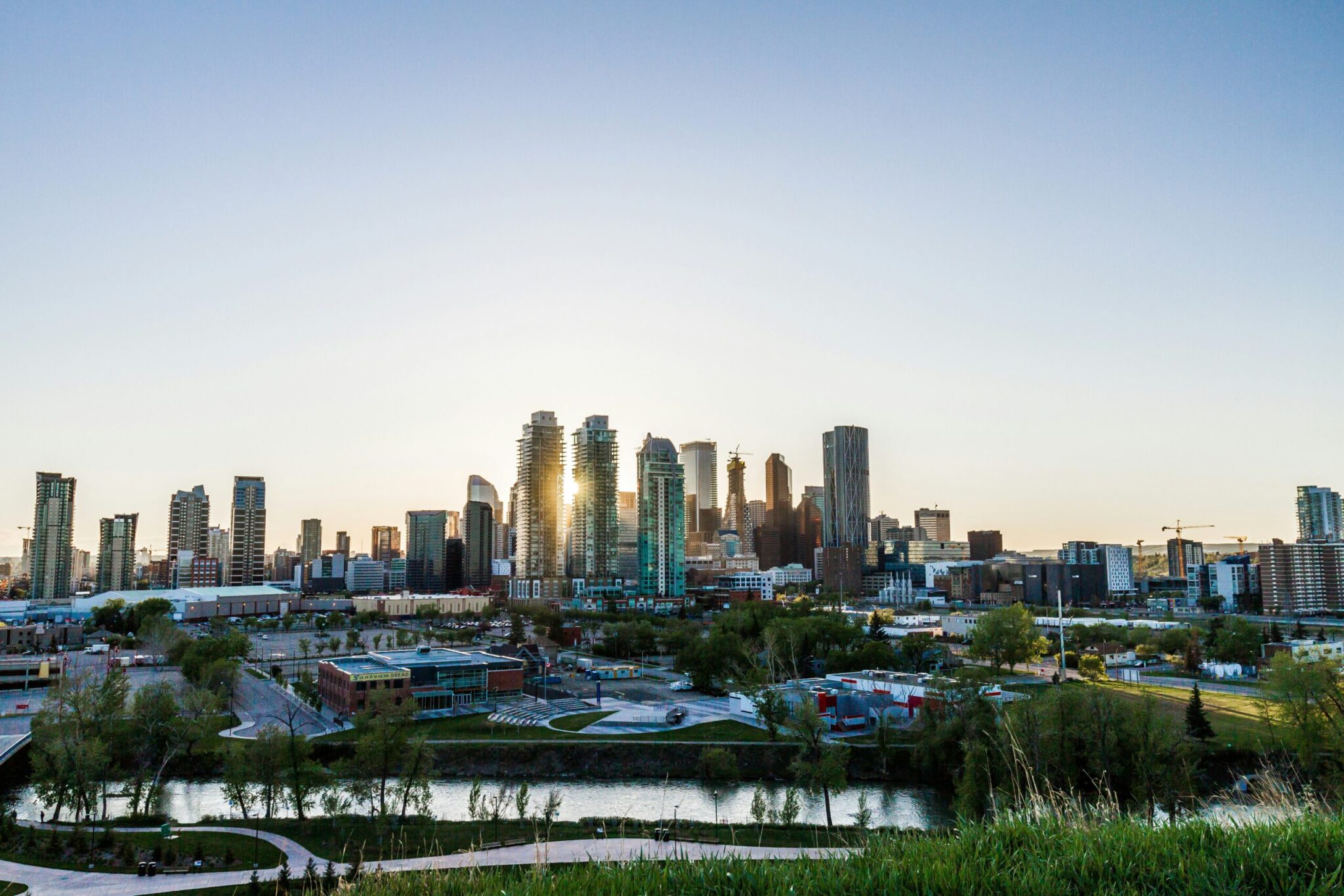 A wide view of a modern city skyline at sunset, with tall glass buildings reflecting sunlight. A river, green park, and various low-rise structures are in the foreground under a clear blue sky.