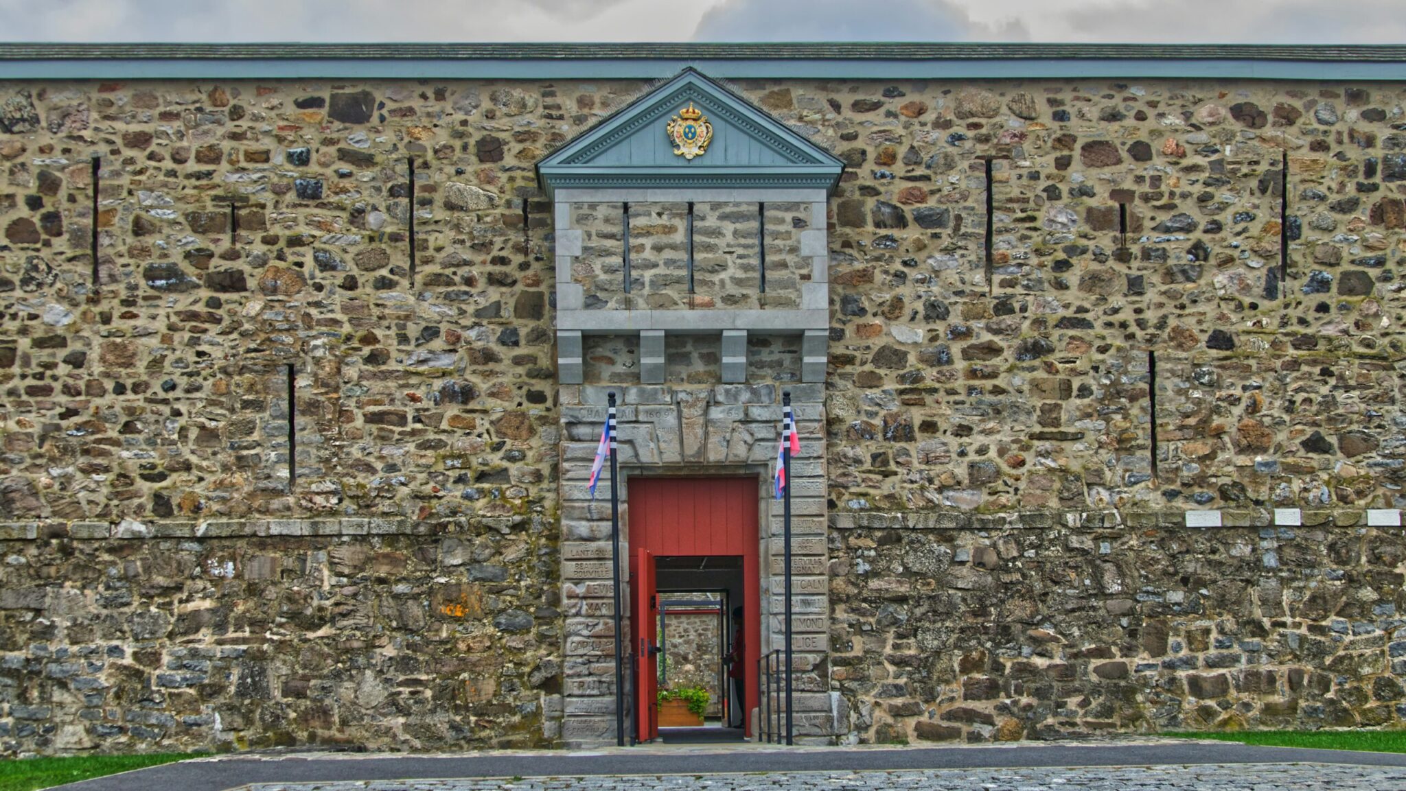 A stone fortress wall with a central arched entrance, flanked by two flagpoles with British flags, a triangular pediment above the door, and a cobblestone path leading inside.