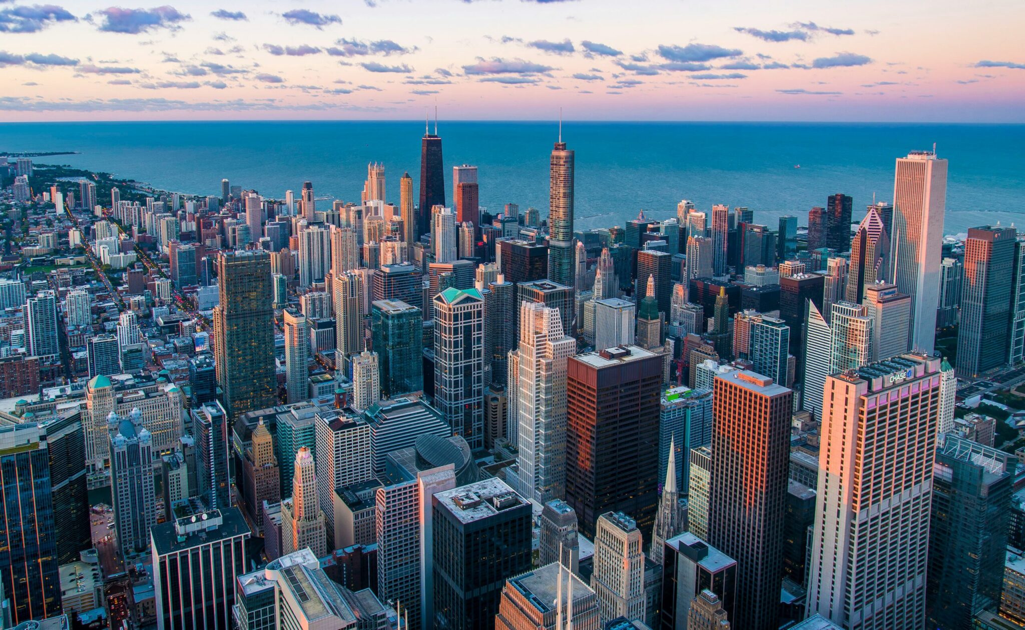 Aerial view of downtown Chicago at sunset, showing tall skyscrapers clustered closely together with Lake Michigan and a lightly clouded sky in the background.