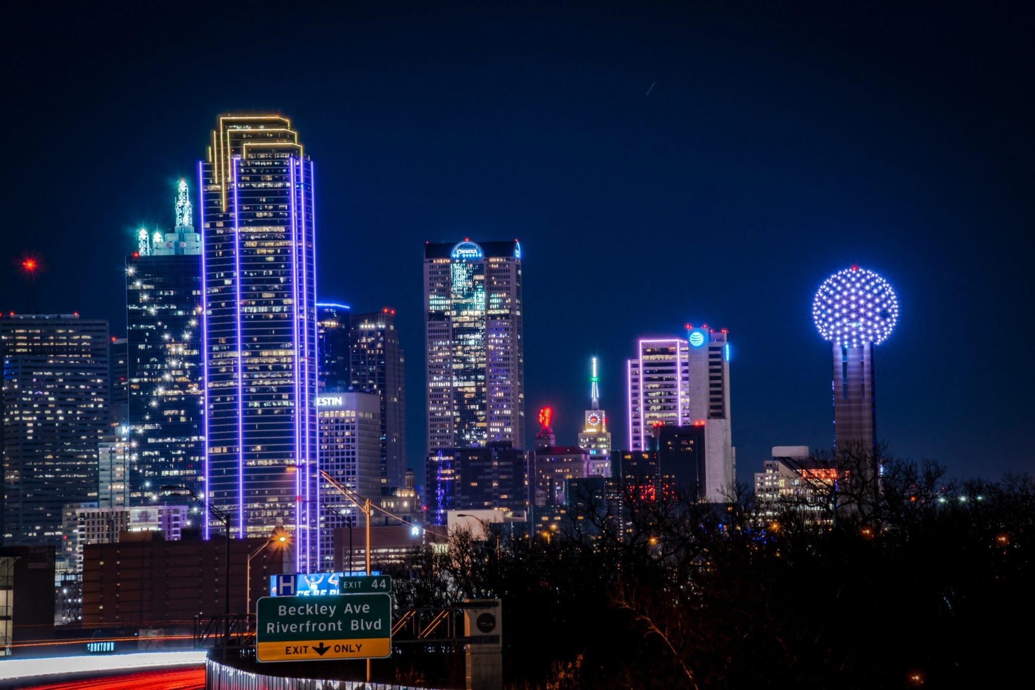 Dallas skyline at night with tall buildings lit in colorful lights, including the Reunion Tower glowing with blue lights. A highway sign for Beckley Ave and Riverfront Blvd is visible in the foreground.