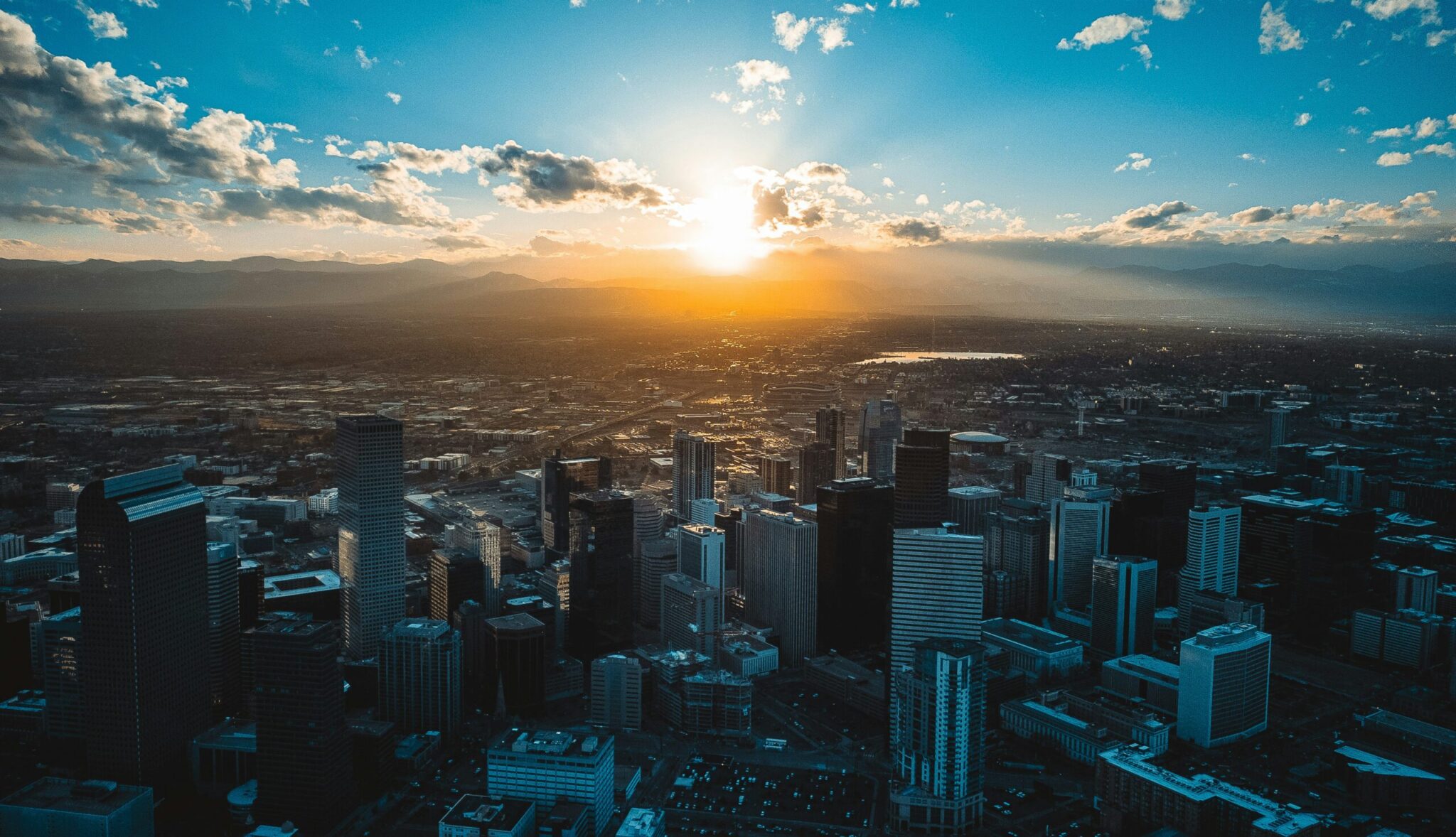 Aerial view of a city skyline at sunset, with tall buildings in the foreground and mountains silhouetted in the distance under a partly cloudy sky. Sunlight streams through the clouds, creating a dramatic effect.