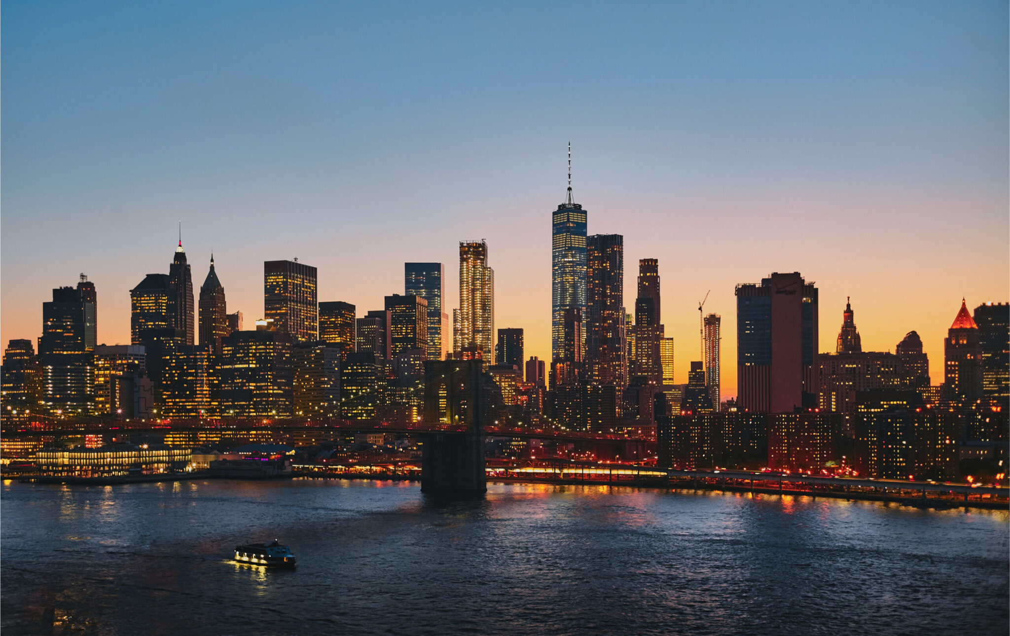 New York City skyline at dusk, with illuminated skyscrapers, including One World Trade Center, reflecting on the East River. A boat is visible on the water in the foreground under a colorful sunset sky.