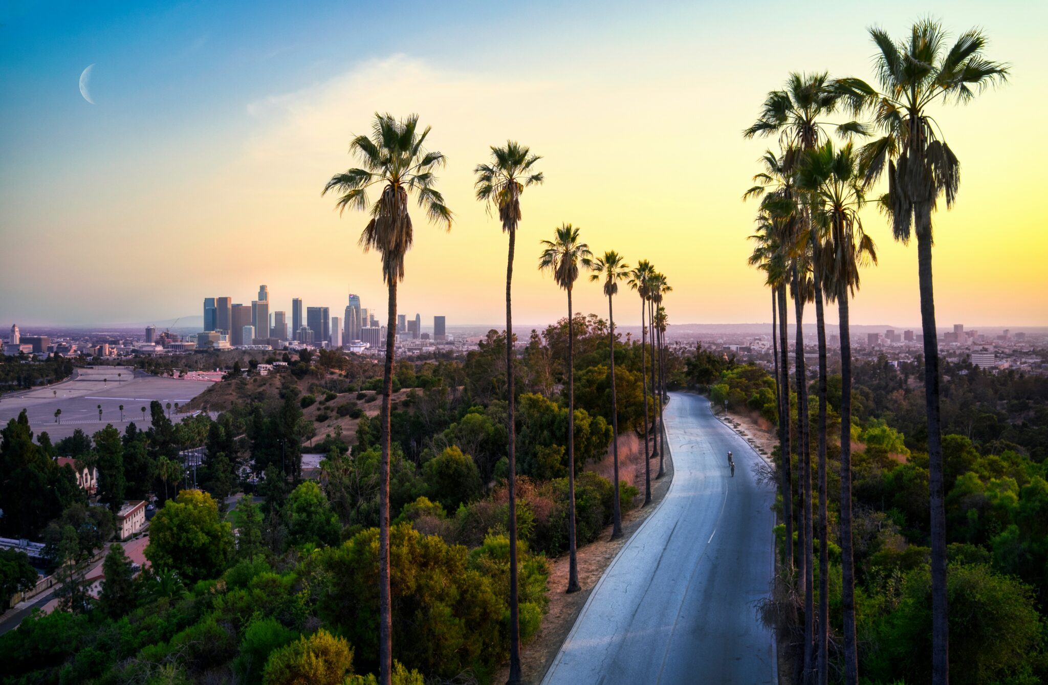 A winding road lined with tall palm trees leads toward downtown Los Angeles at sunset, with the city skyline visible in the distance and a crescent moon in the sky.
