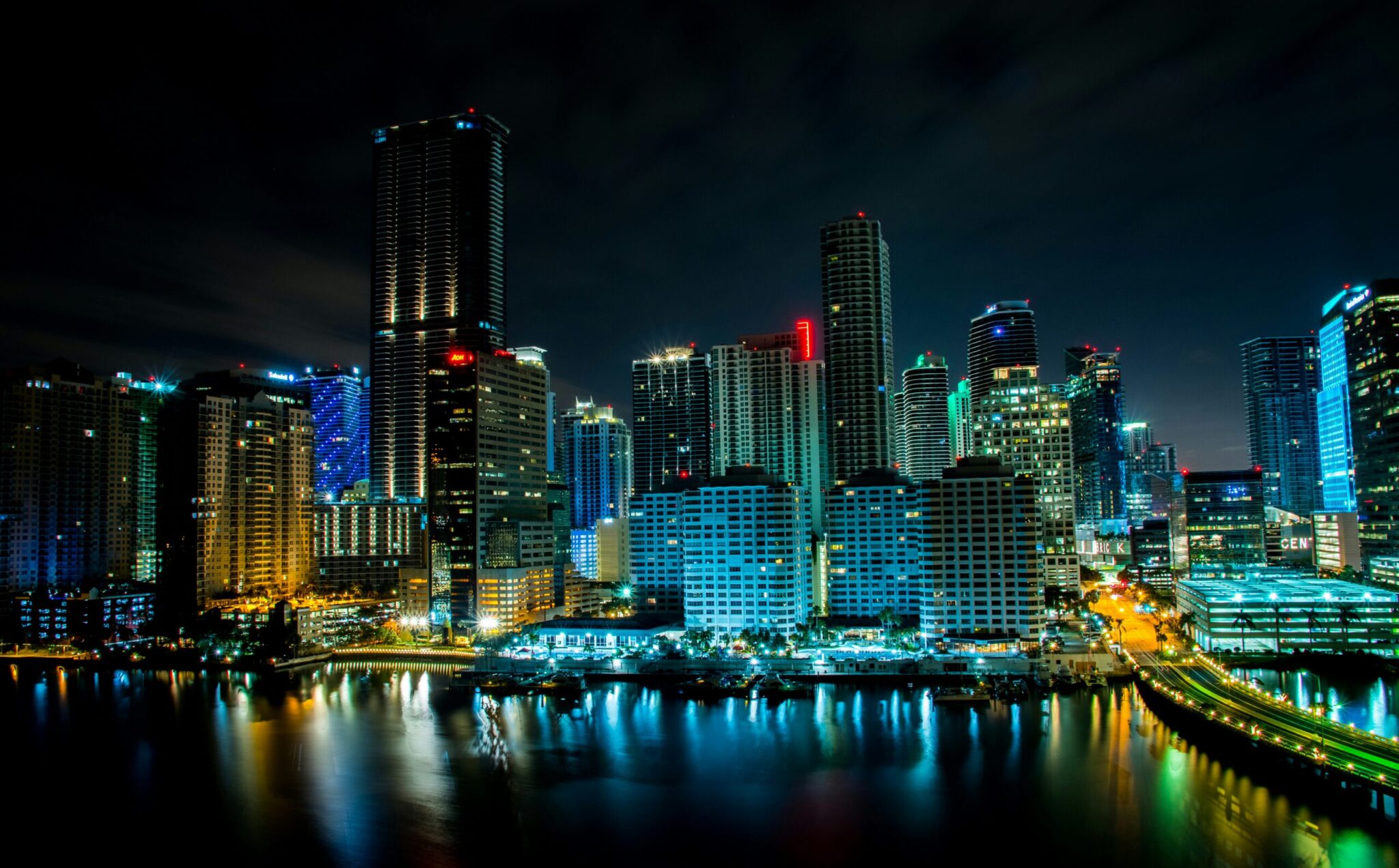 A vibrant city skyline at night with tall, illuminated skyscrapers reflected in the water below. The scene features bright lights, colorful building accents, and a modern urban atmosphere.
