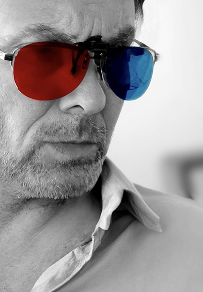 A man with a serious expression wears glasses with one red lens and one blue lens. The photo is mostly black and white except for the colored lenses.