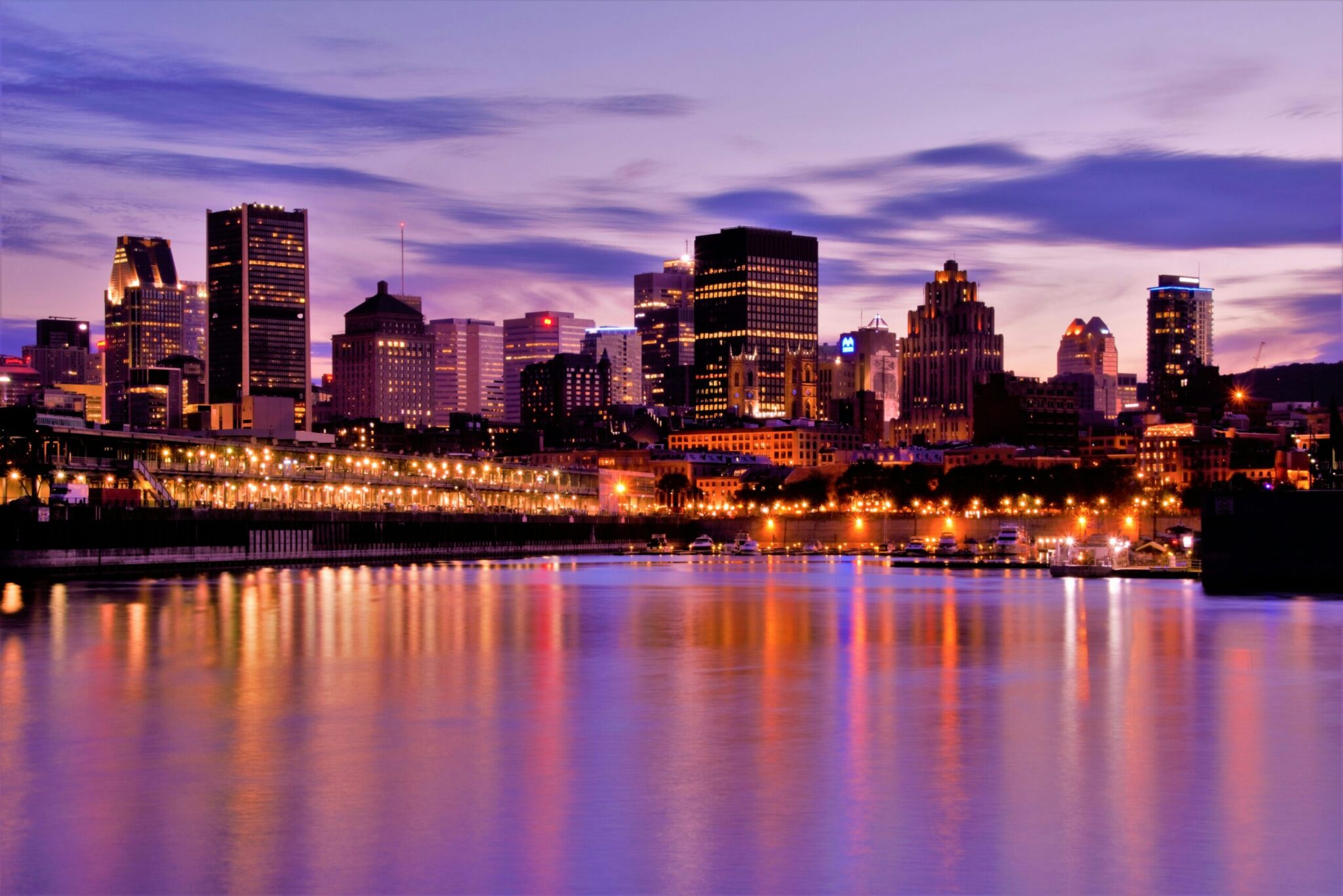 A city skyline at dusk with tall buildings lit up, reflecting colorful lights on the calm water in the foreground under a purple and blue sky.