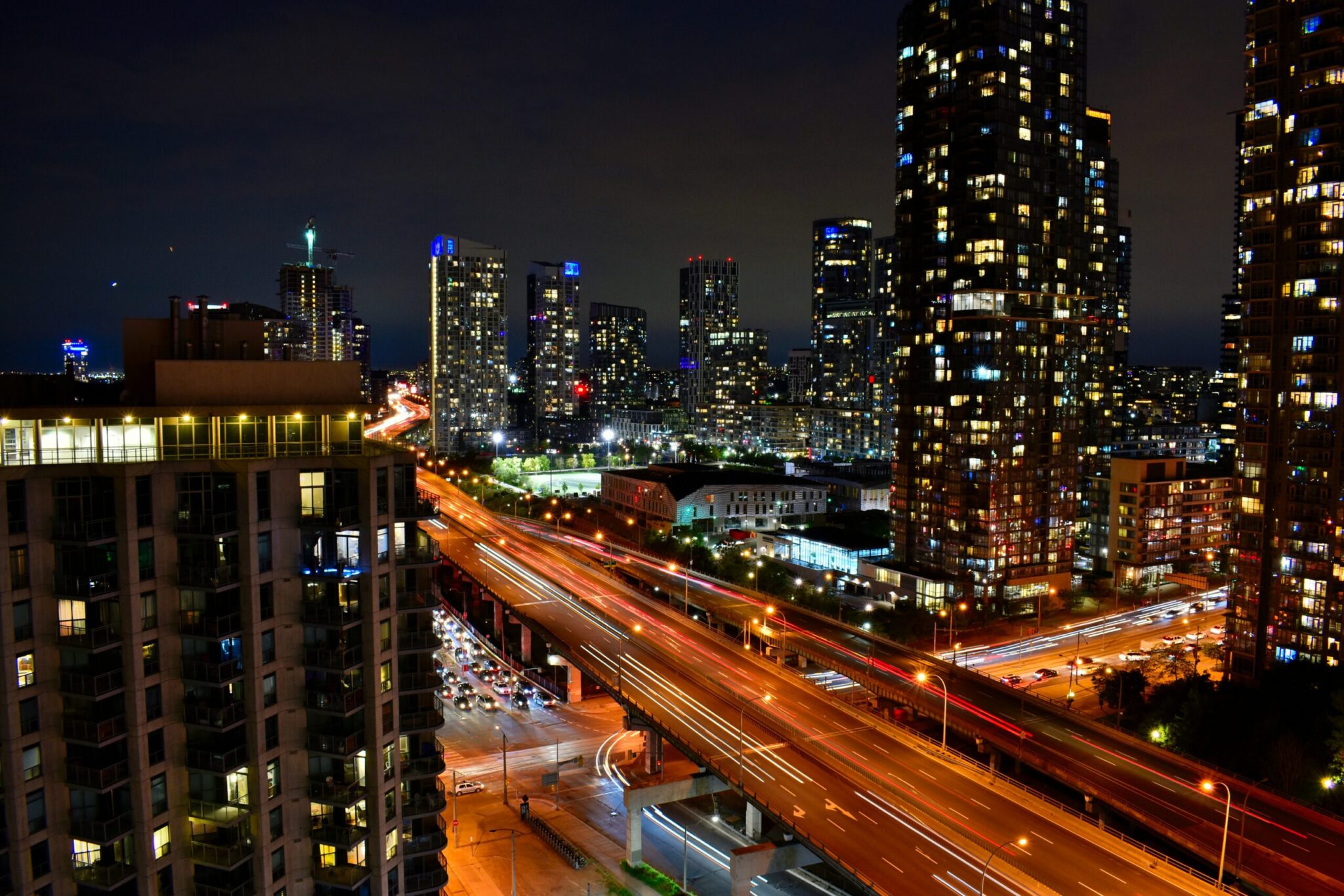 A city skyline at night with tall buildings illuminated by lights, busy highways with streaks of car headlights and taillights, and a vibrant urban atmosphere.