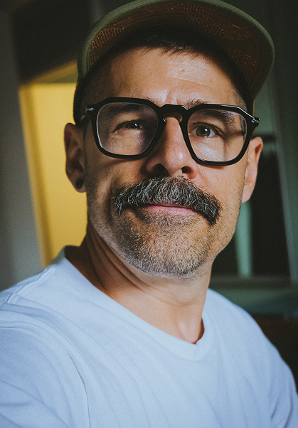 A man with a mustache, beard stubble, large black glasses, a tan cap, and a white shirt looks slightly to the side in a well-lit indoor setting.