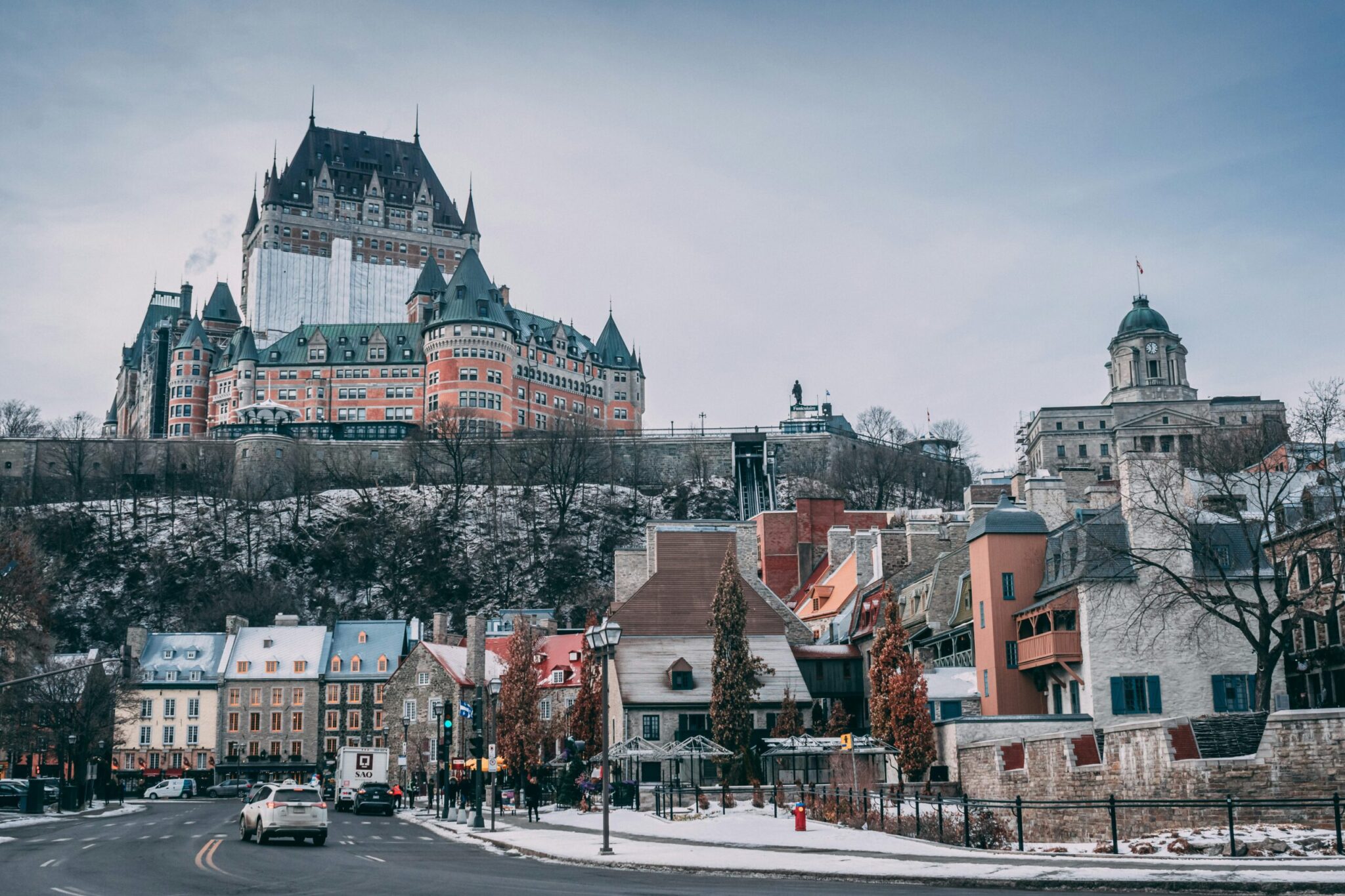 A winter view of Old Quebec City with snow-dusted rooftops, colorful historic buildings, and the grand Château Frontenac hotel towering on a hill under a cloudy sky.