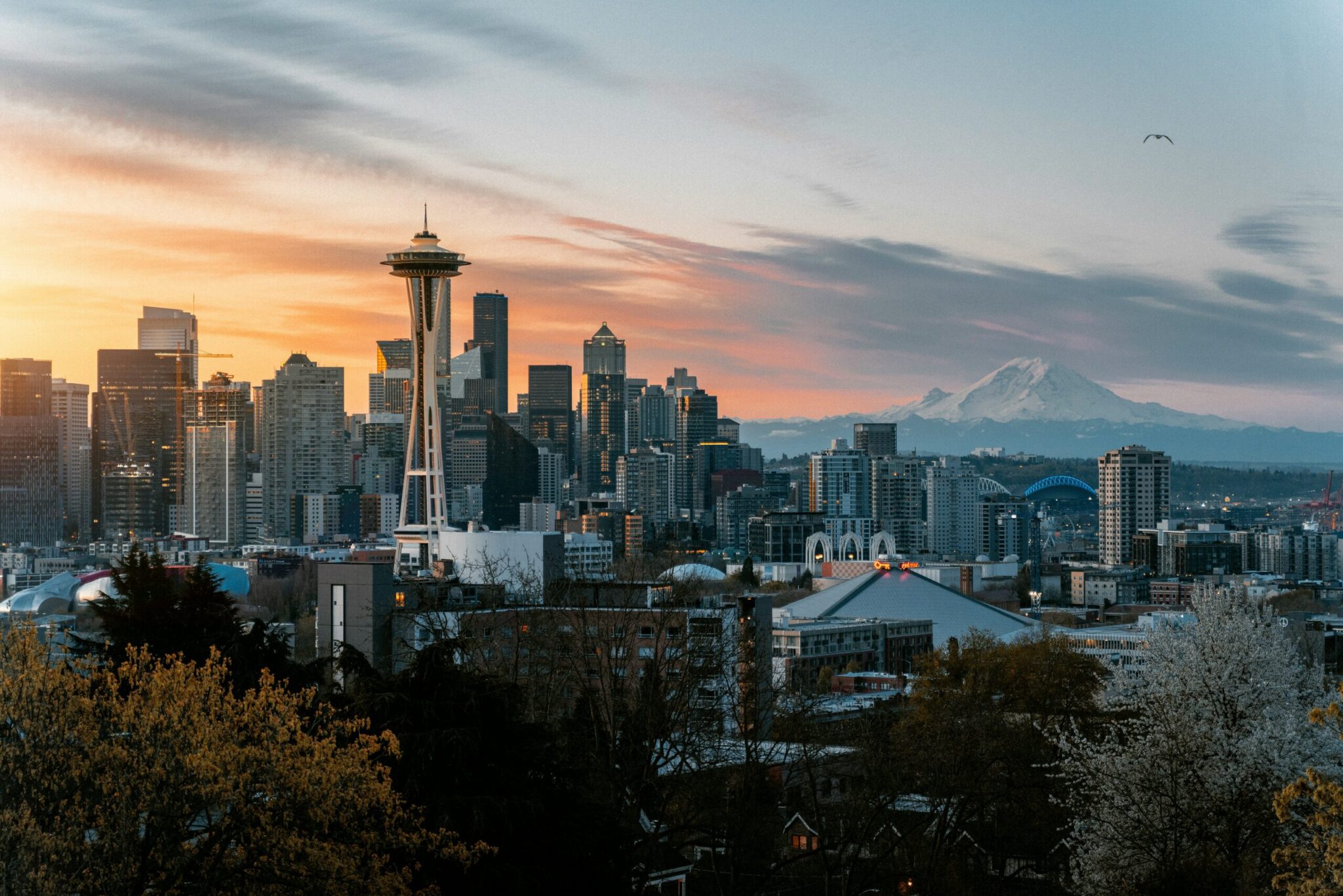 A panoramic view of downtown Seattle at sunset, featuring the Space Needle, city skyscrapers, and Mount Rainier in the background under a partly cloudy sky.