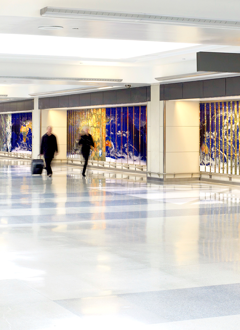 Two people walk through a bright, spacious airport terminal with suitcases. The walls feature large, colorful glass art panels with blue and gold patterns. The floor is shiny and reflective.