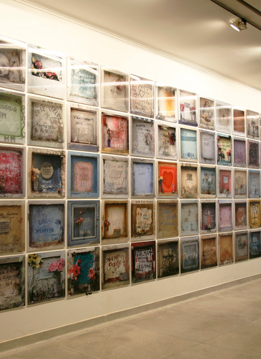 A gallery wall displays a grid of colorful framed photographs, each resembling a memorial plaque or niche with flowers and inscriptions, creating a patchwork effect along the white wall.