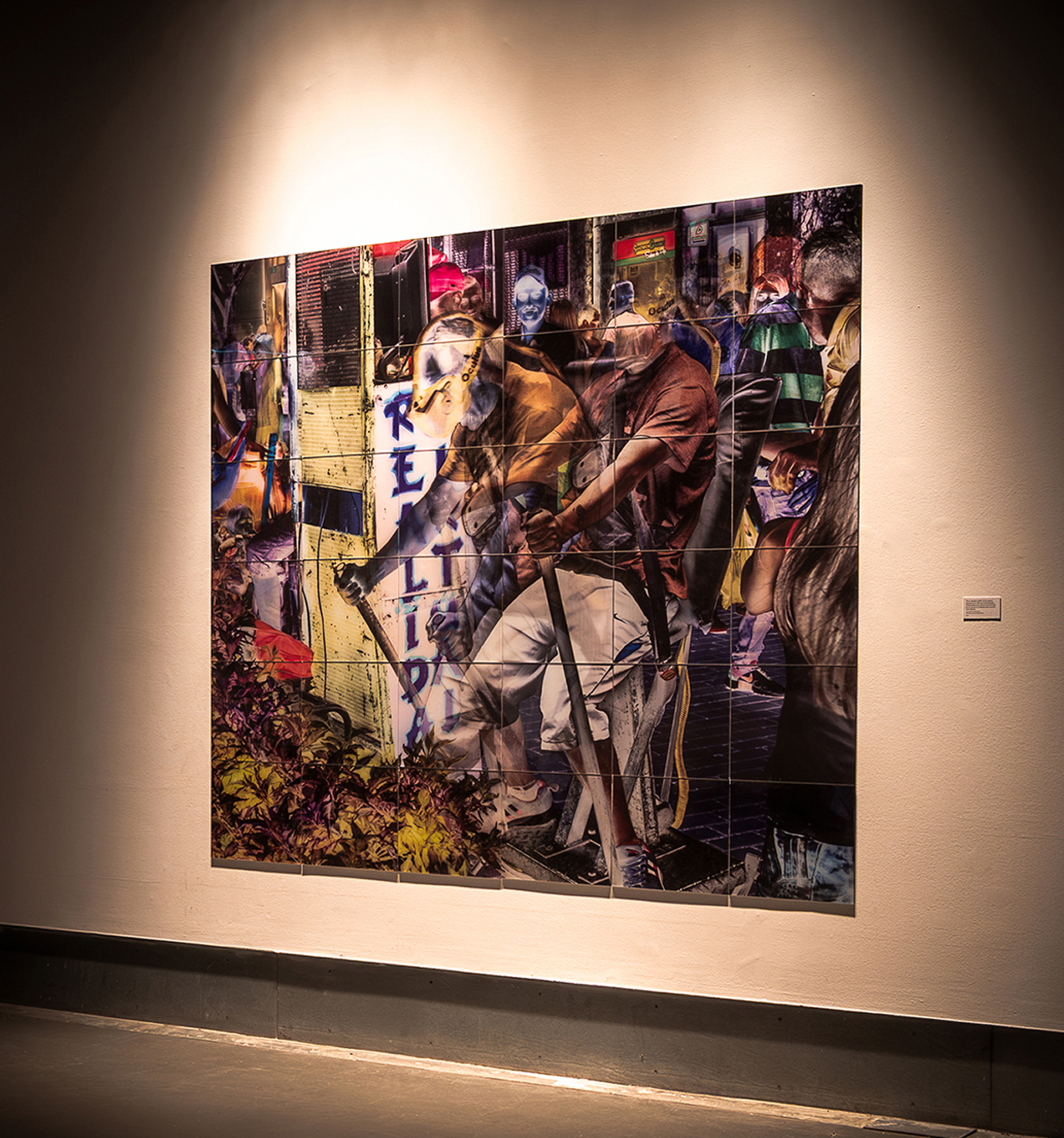 A large, colorful photo collage is displayed on a gallery wall, depicting layered urban scenes with several people, plants, and a vertical sign reading "REALIDAD." The artwork is illuminated by focused lighting.