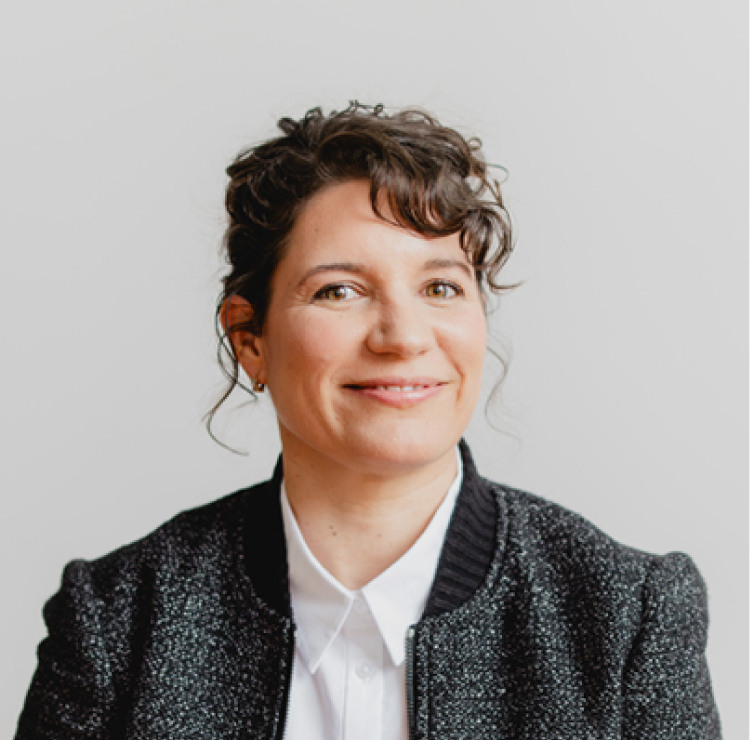 A woman with short, curly dark hair smiles at the camera. She is wearing a textured dark blazer over a white collared shirt and sits against a plain light background.
