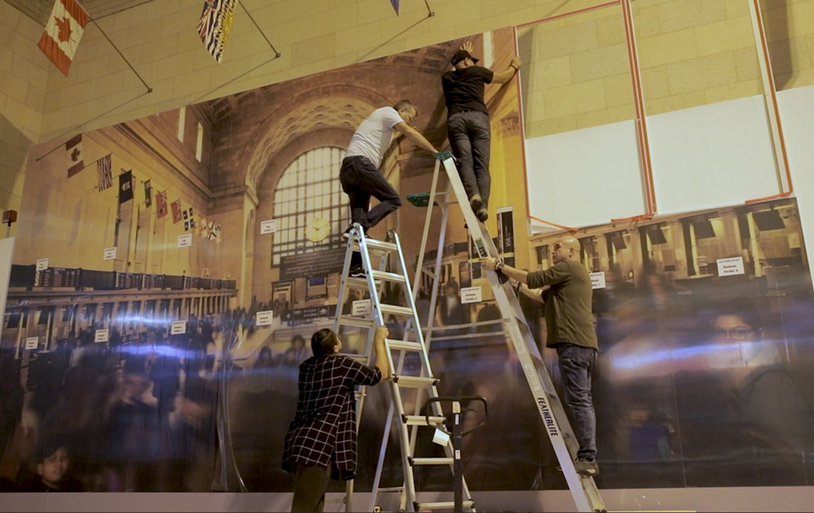 Three people stand on ladders while another steadies a ladder, all working together to hang a large, printed image of a busy train station on an indoor wall. Several flags are visible above them.