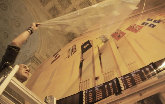 A person standing on a lift raises a clear plastic sheet in front of a large mural featuring international flags and an airport departure board. The ceiling above is ornate, suggesting a grand interior space.
