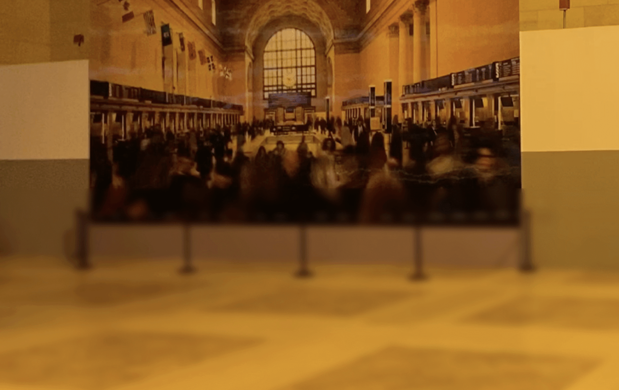A large mural depicting a busy train station scene is displayed on a wall, separated from the viewer by a short barrier. The mural features arched windows and crowds of people inside a grand hall.