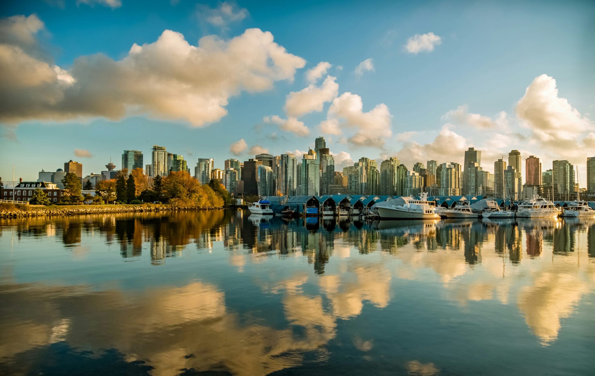 A calm waterfront with boats docked, trees on the left, and a city skyline with tall buildings in the background, all reflected in the water under a partly cloudy sky.