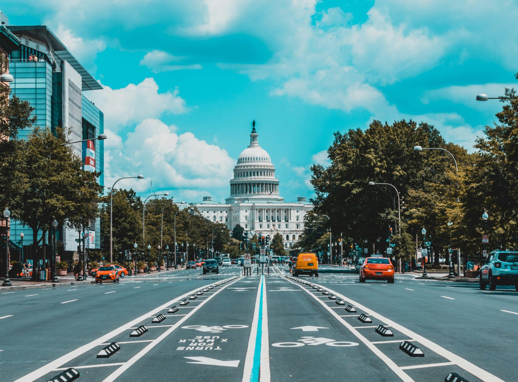 A wide street in Washington, D.C. leads to the U.S. Capitol Building under a partly cloudy sky, with cars, bikes, and trees lining both sides of the road.